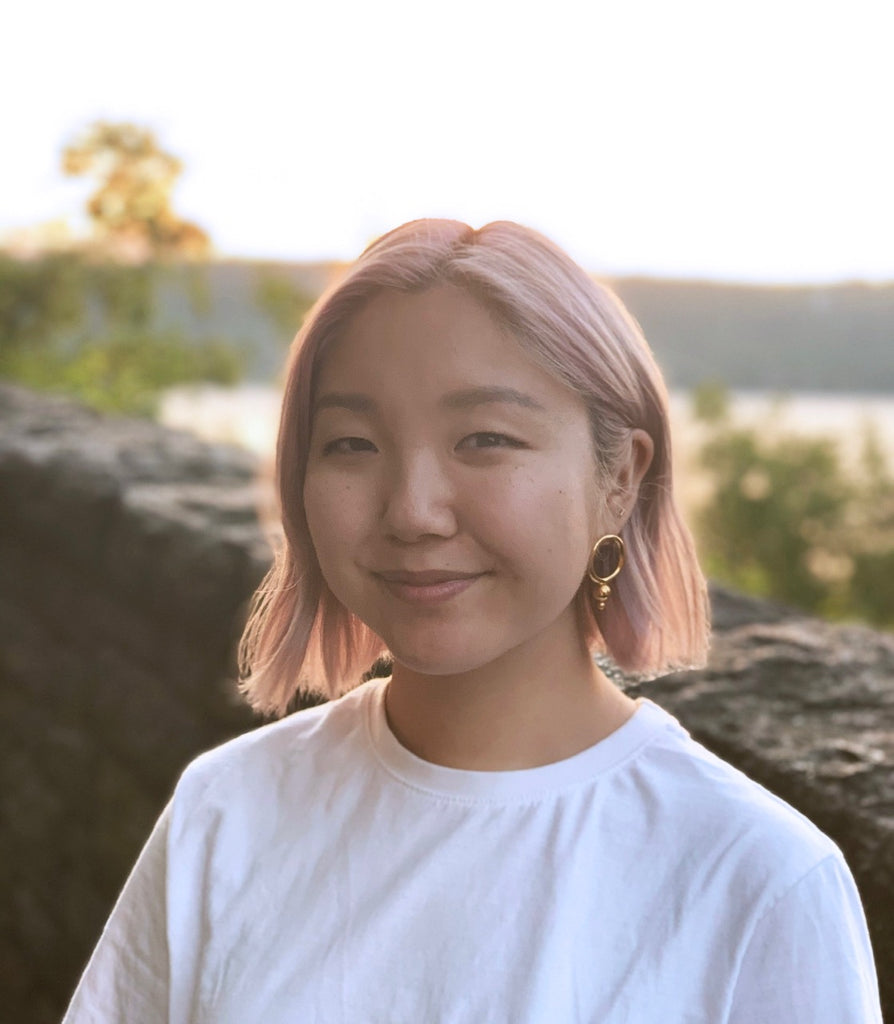 The founder of Halmi in front of a river, during golden hour. She is wearing a white t-shirt, gold earrings and has short, pink hair.