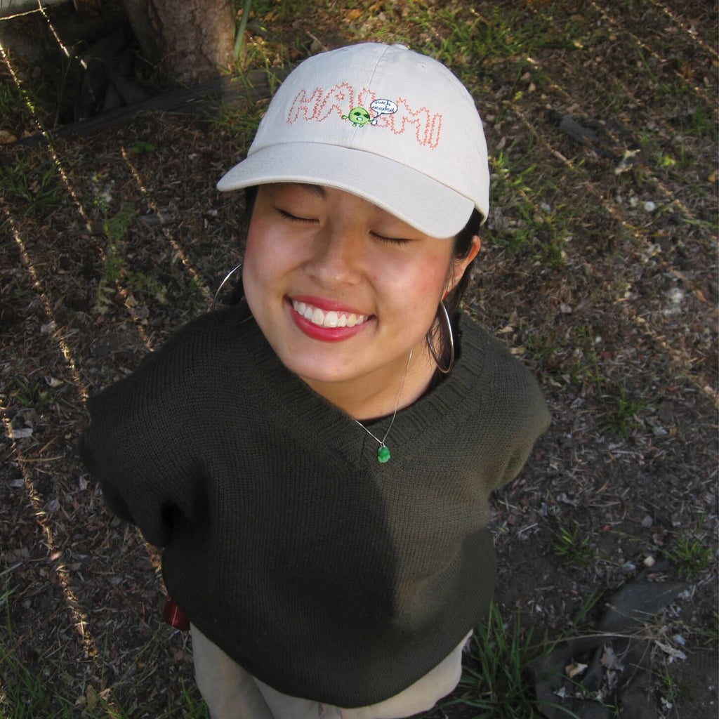 A woman smiling with a stone Halmi cap with embroidered HALMI and green blob saying much needed
