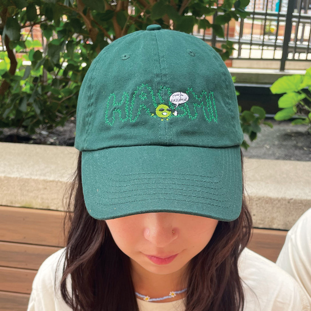 A woman wearing a forest green Halmi cap with embroidered HALMI and green blob saying much needed
